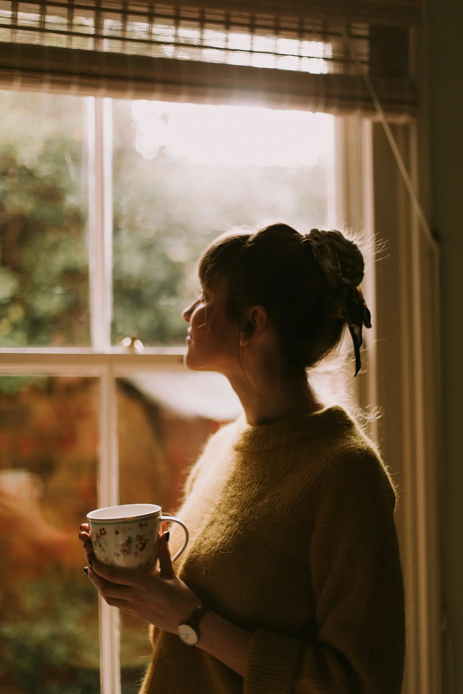 Eine Frau mit einer Tasse in der Hand steht entspannt am Fenster